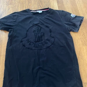 Svart Moncler t-shirt med broderad logga - Svart t-shirt från Moncler i mjuk bomull med stor broderad Moncler-logga på bröstet och klassisk patch på ärmen. Rund halsringning och korta ärmar. Snygg och stilren modell som passar perfekt till jeans eller shorts.