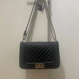 Svart quiltad axelväska med kedja - SKICKA GÄRNA PRISFÖRSLAG! Snygg svart axelväska i skinnimitation med quiltad design och silvrig kedjerem. Väskan har ett stilrent spänne framtill och invändigt fack med dragkedja. Perfekt storlek för det viktigaste och ger en cool vibe till din outfit.