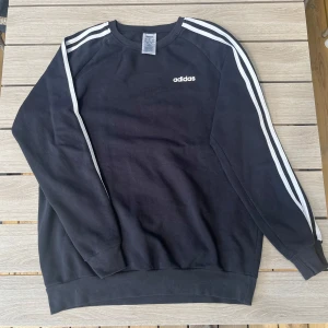 Svart Adidas sweatshirt - Svart sweatshirt från Adidas med klassiska vita ränder längs ärmarna och Adidas-logga på bröstet. 
