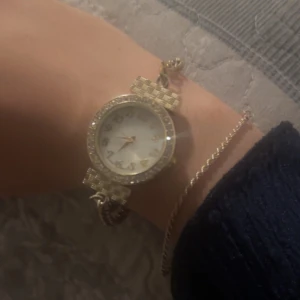 Guldig klocka med glittrande stenar - Säljer en snygg guldig klocka med rund urtavla och glittrande stenar runt hela boetten. Klockan har ett elegant armband i metall med rutmönstrad detalj vid fästena och vit urtavla med guldiga siffror. Perfekt accessoar för att lyfta din stil.