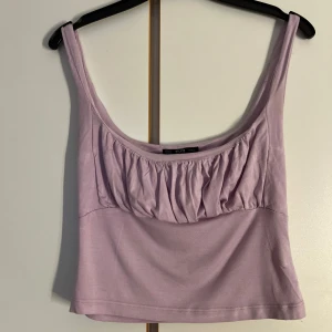 Lila linnetopp från Dilvin  - Supersöt lila linnetopp från Dilvin med snygg rynkad detalj över bysten och breda axelband. Toppen är croppad och har en mjuk, stretchig känsla tack vare materialet i viskos och elastan. Perfekt att styla med jeans eller kjol för en chill look.