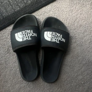 Tofflor från  The North Face - Svarta slides från The North Face med vit logga över foten. Snygg och enkel design med bred rem och öppen tå. Tillverkade i syntetmaterial som är lätta att bära och perfekta för sommaren eller efter träningen.