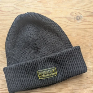 Svart ribbad mössa från Barbour - Svart ribbstickad mössa från Barbour International med uppvikt kant och snyggt gult logomärke framtill. Tillverkad i 60% bomull och 40% akryl för en mjuk och skön känsla. Perfekt accessoar för kalla dagar och enkel att matcha med din stil.