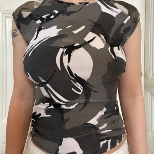 Camo-inspirerad ärmlös topp - Säljer en tight ärmlös topp med camouflagemönster i svart, grått och vitt. Toppen har rund halsringning och figurnära passform, perfekt för dig som gillar edgy och street-inspirerad stil. Materialet är stretchigt och mjukt med mesh vid axlarna.