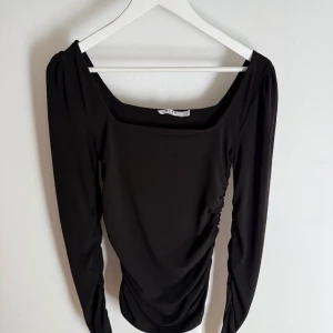 Svart långärmad topp från NA-KD - Stilren svart långärmad topp från NA-KD med fyrkantig ringning och lätt rynkade detaljer längs sidorna. Sitter snyggt och har en smickrande passform. 