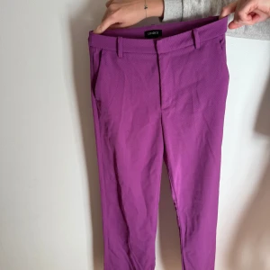 Lila kostymbyxor från Lindex - Snygga lila kostymbyxor från Lindex i modellen Fiona med raka ben och hög midja. Byxorna har hällor för bälte, sidfickor fram och dekorativa fickor bak. Perfekta för dig som vill sticka ut med färg och ändå hålla det stilrent. Otroligt smickrande modell!! Passar mig perfekt i längden som är 167cm. 