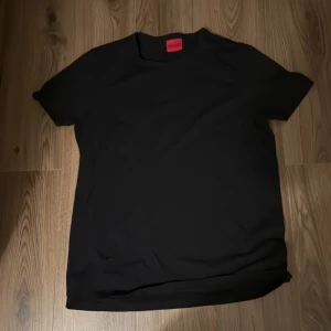 Svart t-shirt från Hugo - En stilren svart t-shirt från Hugo med klassisk passform och rund hals. T-shirten är helsvart utan tryck, med diskret Hugo-logga i nacken och röd etikett i kragen. Perfekt för en clean och enkel look.