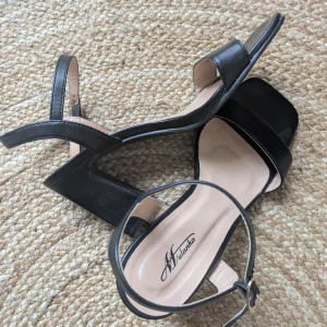 Svarta sandaler med klack från Mylanka - Stilrena svarta sandaler från Mylanka med bred rem över tårna, öppen tå och justerbart spänne vid ankeln. Skorna har fyrkantig tå och stabil blockklack som ger extra höjd. Perfekta för dig som gillar minimalistisk och trendig look. Använt dem 2 ggr på fest 