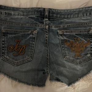 Lowrise jeansshorts med detaljer på bakfickorna - Snygga blå jeansshorts med slitna kanter. Bakfickorna har guldfärgad brodyr. 