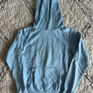 Ljusblå hoodie från Gant - Ljusblå hoodie från Gant med broderad logga på bröstet. Klassisk känguruficka framtill och huva. Tillverkad i mjuk bomullsblandning, perfekt för chill dagar. 