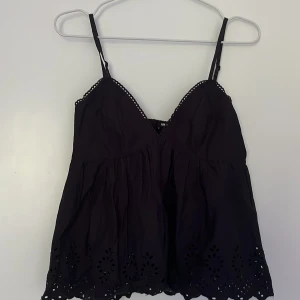 Svart blus från BikBok - Supersöt svart blus från BikBok i storlek XS. Den har smala justerbara axelband, v-ringning och broderade detaljer längs nederkanten. Lätt och luftig modell med volangig känsla och fina hålbroderier som ger en extra touch.