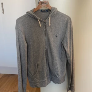 Grå hoodie från Polo Ralph Lauren - Snygg grå hoodie från Polo Ralph Lauren med dragkedja framtill och klassisk huva med vita snören. Tröjan har långa ärmar, två fickor och den ikoniska lilla loggan broderad på bröstet. Perfekt för en chill och stilren look.