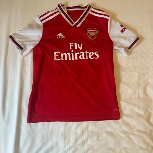Arsenal Adidas matchtröja 2019/20 - Arsenal fotbollströja från Adidas i rött och vitt med klubbmärke och Fly Emirates-tryck på bröstet. Tröjan har korta ärmar, V-ringad krage med ränder och är tillverkad i lätt, ventilerande Climalite-material. Klassisk design med vita axelränder och AFC-detalj nertill.