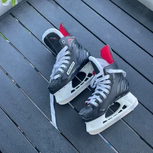 Svarta  hockeyskridskor St 33,5 - Snygga svarta hockeyskridskor från Bauer med vita snören och detaljer i rött. Skridskorna har en robust design med vit sula och tålig ovandel, perfekta för isen. Märket Bauer syns tydligt på sidan och plösen.