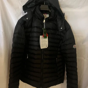 Svart Moncler dunjacka med huva - Snygg svart dunjacka från Moncler med klassisk logga på ärmen. Jackan har huva, dragkedja framtill och två sidofickor med dragkedja. Quiltad design som ger en cool look och håller dig varm. Perfekt för dig som vill ha både stil och funktion under kalla dagar.