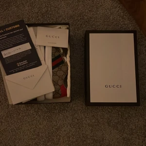 Gucci sneakers (Barn/Kids)  - Snygga Gucci sneakers med klassiskt GG-monogram i beige och brunt, vita snören och vit sula. Skorna har röd och grön rand på sidan samt röd häl på ena skon och svart på den andra. Perfekt för dig som vill sticka ut med lyxig streetstyle.