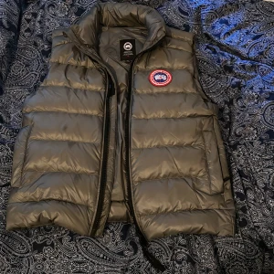 Grå dunväst från Canada Goose - Säljer en grå dunväst från Canada Goose med klassisk logga på bröstet. Västen är quiltad och har hög krage samt dragkedja framtill. Perfekt för lager-på-lager och håller dig varm under kyliga dagar.