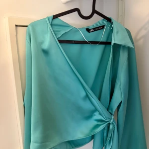 Turkos omlottblus från Zara - Säljer en turkos omlottblus från Zara i glansigt satinliknande material. Blusen har långa ärmar, klassisk krage och knyt i sidan för en snygg passform. Perfekt för dig som vill sticka ut med en färgstark och trendig topp.