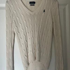 Stickad tröja från Ralph Lauren - Stickad tröja från Ralph Lauren i storlek S. Klassisk v-ringning, långärmad och med mörkblå broderad logga på bröstet. Snygg ribbad kant vid ärmslut och nederkant. På bilderna ser tröjan väldigt beige ut men den är mer vit/kräm färgad som ni ser på dom lite mer närmare bilderna. Tröjan är i nytt skick, säljer på grund av att jag knappt använder den ❤️