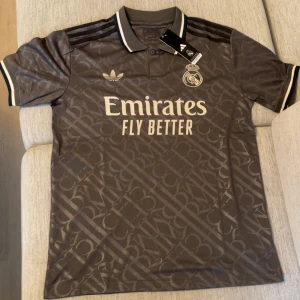 Real Madrid grå fotbollströja - Snygg real Madrid fotbollströja från Adidas med diskret mönster över hela tyget. Tröjan har korta ärmar, klassisk krage och vita detaljer på ärmslut och krage. Stora tryck med klubbmärke och Emirates på bröstet. Perfekt för dig som vill sticka ut på läktaren.