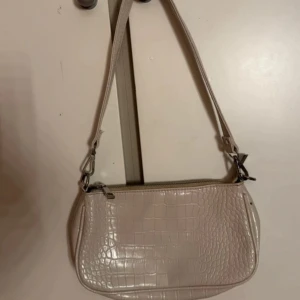 Beige axelväska i crocopräglat skinn - Snygg beige axelväska med crocopräglat mönster och justerbar rem. Väskan har en stilren form och stängs med dragkedja upptill. Perfekt för dig som gillar trendiga accessoarer med lite edge.