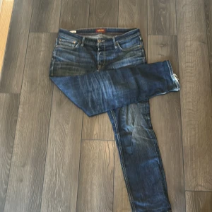 Blå jeans från Jack & Jones - Säljer ett par snygga blå jeans från Jack & Jones med klassisk femficksdesign och slitna detaljer. Jeansen har normal passform och raka ben, med tydliga kontrastsömmar och läderpatch bak i midjan. Perfekta för en avslappnad stil.