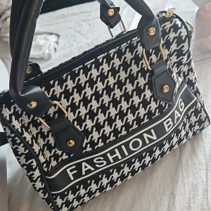 Svartvit handväska med hundtandsmönster - Snygg svartvit handväska med klassiskt hundtandsmönster och texten 'FASHION BAG' framtill. Väskan har svarta handtag med guldfärgade detaljer och dragkedja upptill. Tillverkad i textil och syntetmaterial, perfekt för dig som gillar statement-accessoarer.