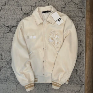 Beige bomberjacka från Zara - Snygg beige bomberjacka från Zara med broderade detaljer på bröstet och ribbade muddar med beigea ränder. Jackan har klassisk krage, tryckknappar och två fickor framtill. Perfekt för dig som vill ha en clean och trendig look.
