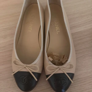 Chanel ballerinaskor i beige och svart - Klassiska ballerinaskor från Chanel i beige skinn med svart tå och rosett framtill. Ikonisk CC-logga på tån och låg platt sula. Perfekt för dig som gillar tidlös och lyxig stil.stl 37 pris 4000kr 