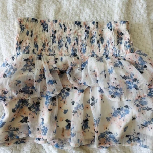 Blommig volangkjol  - Supersöt kort kjol med blommigt mönster i blått och ljusrosa på vit botten. Kjolen har bred smock i midjan och flera lager volanger som ger en luftig och romantisk känsla. Perfekt för varma dagar och somriga outfits.