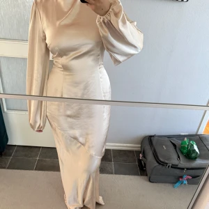 Champagnefärgad långklänning Boohoo - Snygg långklänning från Boohoo i champagnefärgad satinlook. Klänningen har långa ärmar med resår vid handlederna, markerad midja, öppen rygg och knytning i nacken. Materialet är glansigt och känns lyxigt, perfekt för dig som vill sticka ut. Endast använd en gång!