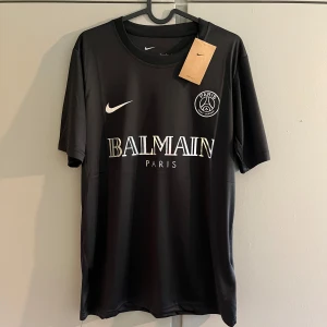 Svart PSG x Balmain fotbollströja Nike - Svart fotbollströja från Nike i samarbete med PSG och Balmain. Tröjan har korta ärmar, rund hals och tryckta loggor i vitt och holografiskt på bröstet. Materialet är lätt och andas, perfekt för träning eller match. Snygg och stilren design med exklusiv känsla.