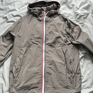 Grå vindjacka från Moncler med huva - Snygg grå vindjacka/regnjacka från Moncler med huva och dragkedja i rött,