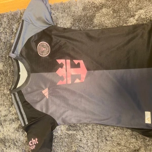 Inter Miami Messi 10 Adidas fotbollströja - Snygg Inter Miami fotbollströja med Messi 10 på ryggen. Tröjan är svart och grå med rosa detaljer, klubbmärke på bröstet och Adidas-logga. Tillverkad i lätt och ventilerande material, perfekt för match eller träning. Storlek S.