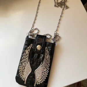 Svart mobilväska med vingar och kedja - Cool svart mobilväska i croc-mönstrat skinnimitation med silverfärgade vingar och strass framtill. Väskan har en lång kedjerem och stängs med magnet knapp. Perfekt för att lyfta din outfit med edgy detaljer.