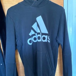 Svart hoodie från Adidas - Svart hoodie från Adidas med stor logga framtill. Klassisk modell med huva och känguruficka.      Pris kan diskuteras 