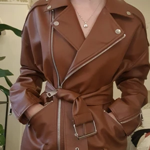 Brun skinnjacka med bälte och dragkedjor - Säljer en brun skinnjacka med coola silvriga dragkedjor och bälte i midjan. Jackan har breda slag, fickor med dragkedja och snygga detaljer på ärmarna. Perfekt för dig som vill ha en edgy och trendig look.