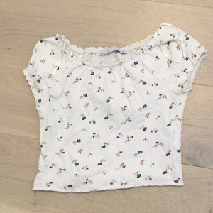 Vit blommig topp från 157 - Supersöt vit topp från 157 med små blommor i olika färger. Modellen har korta ärmar, lätt rynkad halsringning med volangkant.