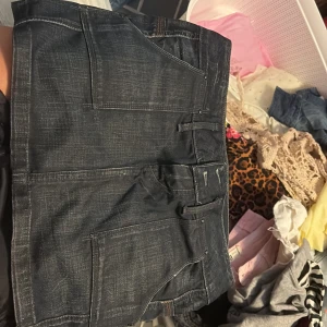 Mörk jeanskjol med fickor och detaljer - Säljer en mörkblå jeanskjol med flera stora fickor, dragkedjor och coola bältesdetaljer i metall. Kjolen har en urban vibe och är perfekt för dig som gillar streetstyle. Snygga kontrastsömmar och slitningar ger en tuff look.