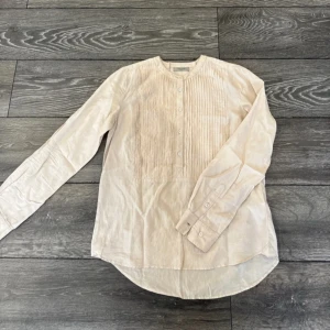 Beige henley-skjorta från HunkyDory - Snygg beige henley-skjorta från HunkyDory med rund halsringning och knappar framtill. Skjortan har lång ärm och coola veckade detaljer på bröstet. Perfekt för en chill och stilren look.