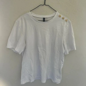 Vit t-shirt med guldknappar - Vit t-shirt från H&M med rund halsringning och tre dekorativa guldknappar på ena axeln. T-shirten har korta ärmar och är tillverkad i mjuk bomull, perfekt för en clean och enkel stil. Endast använd en gång.