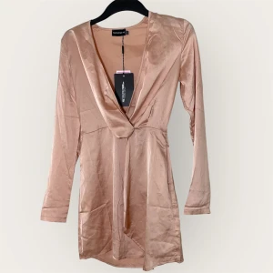 Champagnefärgad klänning PLT - Snygg kort klänning i champagnefärgad satin från PrettyLittleThing. Djup v-ringning, omlott framtill och långa ärmar. Materialet har en lyxig glans och klänningen har dragkedja i sidan för enkel påklädning. Perfekt för dig som vill sticka ut.