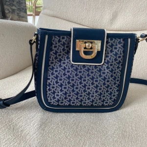Blå axelväska från DKNY med monogram - Snygg blå axelväska från DKNY med justerbar rem och guldigt spänne framtill. Väskan har ett stilrent monogrammönster i blått och vitt, detaljer i skinn och dragkedja på insidan. Perfekt storlek för det viktigaste och en riktigt trendig accessoar.