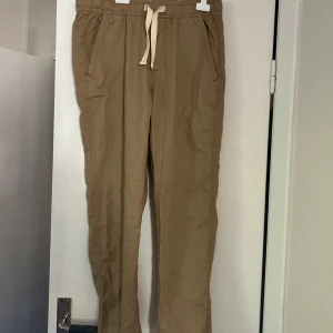 Beiga chinos med snörning - Snygga beiga chinos från Zara med dragsko i midjan och raka ben. Byxorna har sidfickor fram och två bakfickor. Perfekta för en avslappnad men stilren look. Tillverkade i bomull för en skön känsla. Aldrig använda.