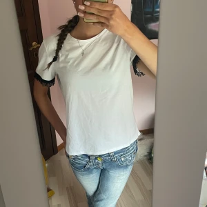 Vit t-shirt med spetsdetalj på ärmar - Vit t-shirt i bomull med korta ärmar och svart spetskant som ger en snygg kontrast. Modellen är rak och har en klassisk rund halsringning. Perfekt för dig som gillar enkla plagg med en twist. Är i nyskick helt oanvänd köpt på zara