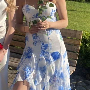 Vit och blå blommig långklänning - Superfin långklänning med vita och blå blommor, smala axelband och volangdetaljer längs kjolen. Klänningen har en feminin passform med markerad midja och lätt utsvängd kjol. Perfekt för somriga tillfällen. Den har även en slits vid ena benet. De är dragkedja där bak❤️