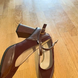 Bruna Mary Jane pumps med klack - Snygga bruna Mary Jane pumps med fyrkantig tå och bred klack. Skorna har en mix av mocka och skinn, dekorativa broguedetaljer och justerbart spänne över foten. Perfekta för dig som gillar retrovibbar och vill sticka ut med en klassisk men cool look.