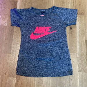 Nike gråmelerad t-shirt med rosa logo - Nike t-shirt i gråmelerat med korta ärmar och stor rosa Nike-logga framtill. Skön och sportig passform, perfekt för träning eller chill. Materialet känns lätt och andas bra. Snygg och enkel design som funkar till det mesta.