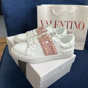 Valentino Rockstud sneakers vit/rosa - Snygga Valentino Rockstud sneakers i vitt skinn med ljusrosa band och ikoniska nitar. Klassisk rund tå, platt sula och vita snören. Märkeslogga på plösen och insidan. Perfekta för dig som vill sticka ut med exklusiva detaljer.