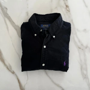 Svart skjorta från Polo Ralph Lauren - Klassisk svart skjorta från Polo Ralph Lauren med smal passform. Skjortan har långa ärmar, vita knappar och en broderad lila logga på bröstet. Tillverkad i bomull och passar perfekt till dig som gillar stilrena plagg.